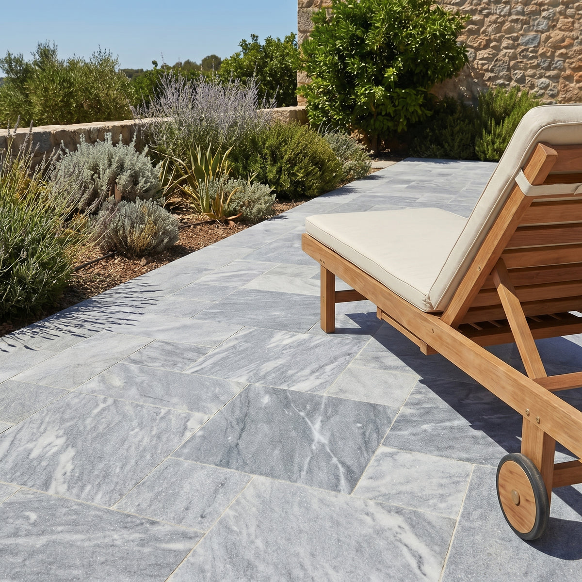 Wooden lounge chair with white cushion on a stone patio with garden in the background. Bluestone tumbled pavers