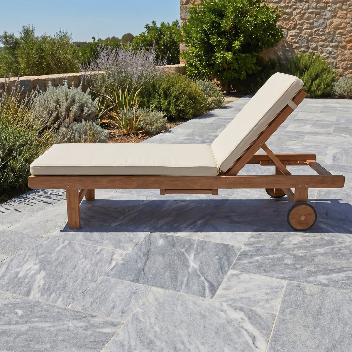 Wooden lounge chair with beige cushions on a stone patio with plants in the background. Bluestone tumbled pavers