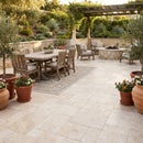 Outdoor patio with dining table, chairs, and fire pit area surrounded by potted plants. Travertine Miletos Ivory Tumbled Pavers