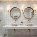 Bathroom with two round mirrors, gold frames, and white sinks on a marble-patterned wall. Palia White Dolomite Dimensional Mosaic Tile Stella Design on 12" x 12" Mesh